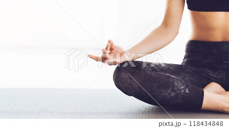 Cropped of woman is sitting cross-legged on a yoga mat in a lotus position, demonstrating flexibility and focus. She appears relaxed and concentrated, with hands resting on her knees, copy space 118434848