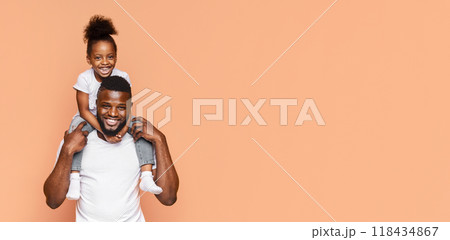 Happy Black father and his daughter posing for a photo together. The father is holding his daughter on his shoulders and they are both smiling widely. The background is a simple peach color Happy Black father and his daughter posing for a photo together. The father is holding his daughter on his shoulders and they are both smiling widely. The background is a simple peach color 118434867
