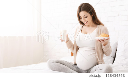 Young pregnant woman having brunch of milk and croissant, sitting on bed and looking at belly, free space Young pregnant woman having brunch of milk and croissant, sitting on bed and looking at belly, free space 118434868
