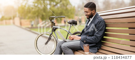 An African American man works on his laptop while sitting on a park bench, his bicycle resting nearby. The setting is peaceful and productive, combining work with the outdoors. 118434877