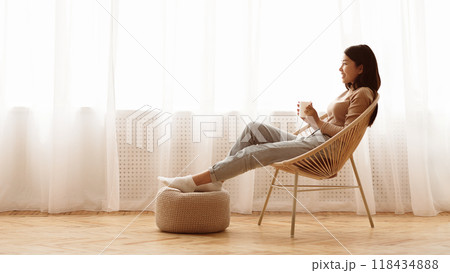 A woman in a brown sweater sits in a wicker chair with her feet up on an ottoman while looking out a window. A woman in a brown sweater sits in a wicker chair with her feet up on an ottoman while looking out a window. 118434888