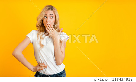 A young woman with long blonde hair stands against a bright yellow background, looking surprised and covering her mouth with her hand. She is wearing a white t-shirt, copy space 118434918