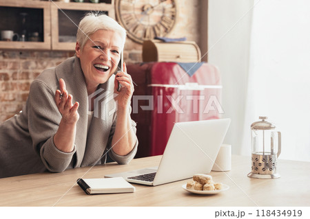 A senior woman with short grey hair is sitting at a kitchen table, smiling and talking on a cell phone. There is a laptop in front of her, along with a notebook A senior woman with short grey hair is sitting at a kitchen table, smiling and talking on a cell phone. There is a laptop in front of her, along with a notebook 118434919
