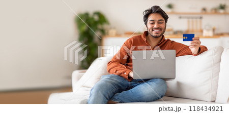 A young Indian man is sitting comfortably on a couch, smiling as he uses his laptop while holding a credit card. The cozy living space has a relaxed atmosphere, perfect for online shopping. 118434921