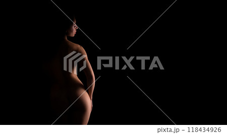 A nude woman is standing in the darkness, illuminated by a faint light source, casting shadows around her. Her figure is silhouetted against the black background. A nude woman is standing in the darkness, illuminated by a faint light source, casting shadows around her. Her figure is silhouetted against the black background. 118434926