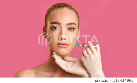 A woman with blonde hair is receiving a facial injection from a gloved hand. She is looking directly at the camera and has a neutral expression on her face. The background is a bright pink color. 118434960