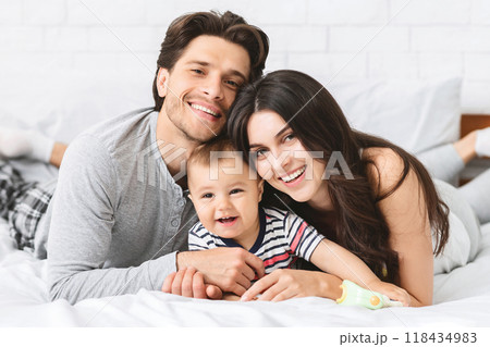 A smiling family of three lies on a bed together. The parents are both looking at the camera, while the baby is smiling at someone out of frame. The family looks relaxed and happy. 118434983
