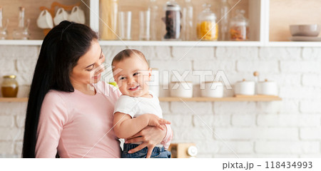 A woman standing, holding a baby in her arms. The woman face is visible and she appears to be looking down at the baby with care and tenderness, copy space A woman standing, holding a baby in her arms. The woman face is visible and she appears to be looking down at the baby with care and tenderness, copy space 118434993