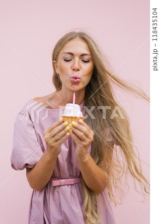 Young caucasian woman is blowing out birthday candle on a pink cupcake. She is wearing a pink dress and long blonde hair 118435104