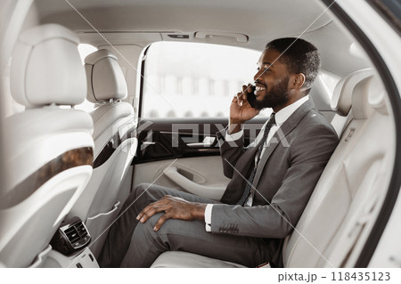 African American businessman in a suit is sitting in the backseat of a car, talking on his phone with a smile on his face. He is dressed in a gray suit, white shirt, and a dark tie African American businessman in a suit is sitting in the backseat of a car, talking on his phone with a smile on his face. He is dressed in a gray suit, white shirt, and a dark tie 118435123