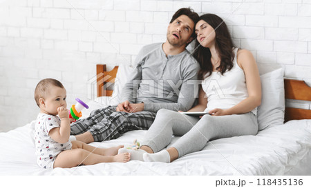 A young couple sits exhausted on a bed, while their baby plays with a toy on the white sheets. The father looks tired and leans back, while the mother seems to be nodding off. 118435136