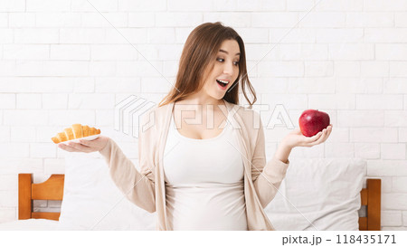 Emotional pregnant woman choosing between red apple, comparing fresh fruit and croissant, sitting on bed Emotional pregnant woman choosing between red apple, comparing fresh fruit and croissant, sitting on bed 118435171
