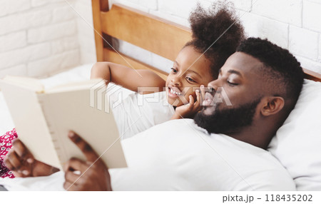 African American father lies in bed reading a book to his little daughter. She is listening intently and smiling. The room is decorated with a white brick wall and a wooden bed frame. African American father lies in bed reading a book to his little daughter. She is listening intently and smiling. The room is decorated with a white brick wall and a wooden bed frame. 118435202