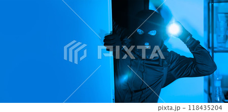 A person dressed in dark clothing and a ski mask peers into a dimly lit room while holding a flashlight, suggesting an illicit entry at night. The atmosphere is tense and foreboding. A person dressed in dark clothing and a ski mask peers into a dimly lit room while holding a flashlight, suggesting an illicit entry at night. The atmosphere is tense and foreboding. 118435204