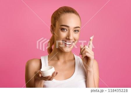 A young woman, with blonde hair pulled back, smiles at the camera as she applies a white cream to her finger. She is holding an empty container in her other hand. The background is a vibrant pink. 118435228