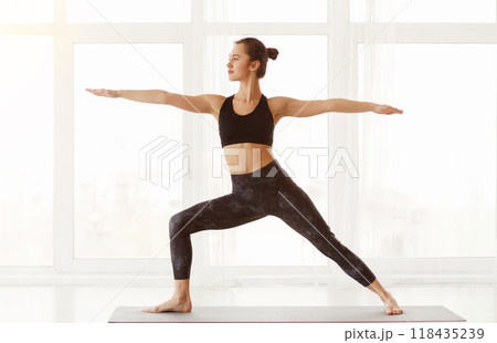 A young woman stands on a yoga mat, arms extended in Warrior II pose, exuding tranquility and focus in a sunlit, spacious studio room 118435239