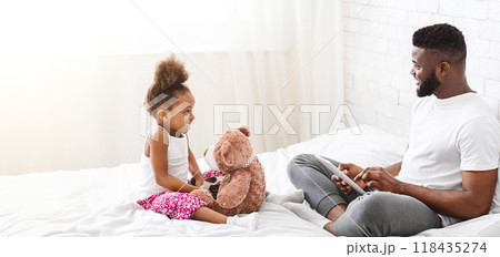 Black father and his little daughter are sitting on a bed, both smiling. The daughter is holding a stuffed bear, while the father is looking at his daughter with a tablet in his lap Black father and his little daughter are sitting on a bed, both smiling. The daughter is holding a stuffed bear, while the father is looking at his daughter with a tablet in his lap 118435274