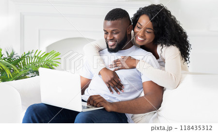 A couple relaxes on a white couch while the man uses a laptop. The woman has her arm around him. 118435312