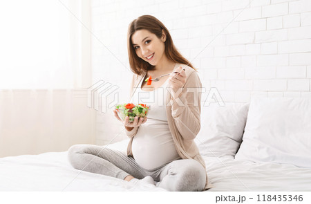 Care about unborn child. Happy pregnant woman eating natural vegetable salad, sitting on bed, panorama with free space Care about unborn child. Happy pregnant woman eating natural vegetable salad, sitting on bed, panorama with free space 118435346