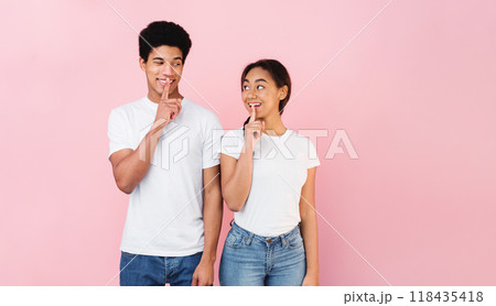 Shhh, it's our secret. Teen couple holding fingers on lips and looking at each other, pink studio background 118435418