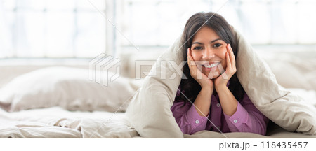 A cheerful Arabic woman sits on a bed, smiling while wrapped in a soft blanket. Bright light filters through the window, enhancing the cozy atmosphere of this serene indoor setting. A cheerful Arabic woman sits on a bed, smiling while wrapped in a soft blanket. Bright light filters through the window, enhancing the cozy atmosphere of this serene indoor setting. 118435457