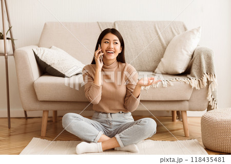 A young woman sits on the floor of a living room, smiling while talking on her phone. 118435481