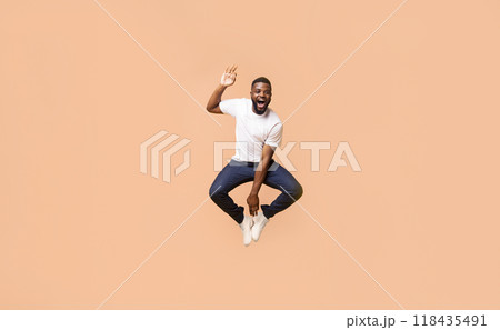 A Black man is jumping in mid-air with his arms raised and an ecstatic expression on his face. He is wearing a white t-shirt and blue jeans. The background is a peach color. A Black man is jumping in mid-air with his arms raised and an ecstatic expression on his face. He is wearing a white t-shirt and blue jeans. The background is a peach color. 118435491