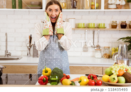 A woman wearing an apron and oven mitts stands in a kitchen with fresh produce and a blender, looking excited. 118435493