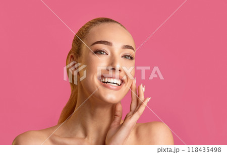 A young woman with long blonde hair smiles broadly, her hand resting on her cheek, against a bright pink backdrop. Her clear skin and white teeth make for a beautiful and radiant image. 118435498