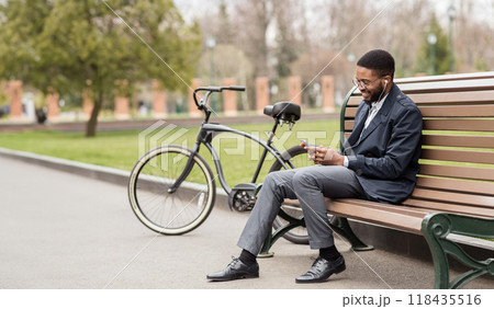 Black businessman is talking on his phone while sitting on a bench in the park, with a bicycle nearby. The atmosphere is relaxed and efficient, set in a modern outdoor environment. 118435516