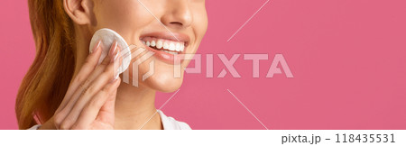 A close-up image of a woman with long brown hair smiling while applying a cotton pad to her cheek in front of a solid pink background. The womans eyes are not visible, cropped, blank space 118435531
