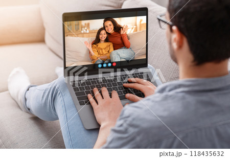 A man sits on a couch, wearing casual clothing and glasses, using a laptop to video chat with his wife and daughter. The woman and girl are sitting together, waving at the man. A man sits on a couch, wearing casual clothing and glasses, using a laptop to video chat with his wife and daughter. The woman and girl are sitting together, waving at the man. 118435632
