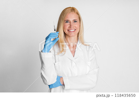 A blonde lady doctor is smiling while holding a syringe in her right hand, wearing blue latex gloves. She is wearing a white lab coat and has her arms crossed. The background is a plain white wall. A blonde lady doctor is smiling while holding a syringe in her right hand, wearing blue latex gloves. She is wearing a white lab coat and has her arms crossed. The background is a plain white wall. 118435650