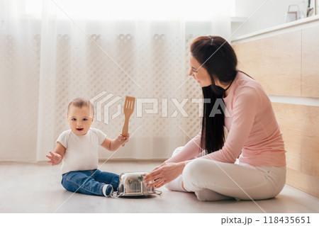 A woman is kneeling down next to a baby who is holding a spatula. The woman is likely interacting with the baby and engaging in a cooking or baking activity together. A woman is kneeling down next to a baby who is holding a spatula. The woman is likely interacting with the baby and engaging in a cooking or baking activity together. 118435651
