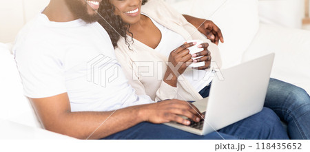 A couple is relaxing on a sofa, the man is using a laptop while the woman holds a mug in her hands. A couple is relaxing on a sofa, the man is using a laptop while the woman holds a mug in her hands. 118435652