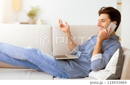 A man sits on a couch with a laptop in his lap and talks on a phone with a smile on his face. 118435664