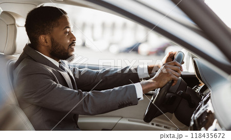 A professional black man wearing a suit and tie is driving a car. He is looking out the window to the right of the frame. The sun is shining brightly, making the inside of the car look bright. 118435672