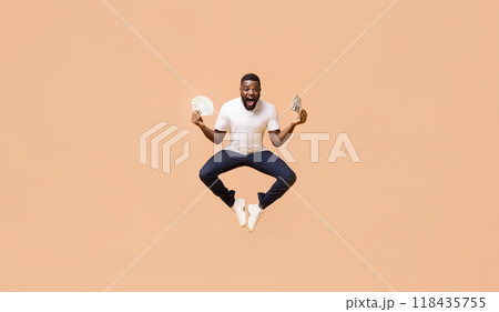 African American young man in a white t-shirt and dark jeans is jumping in the air, with his arms outstretched, holding stacks of money in each hand. The background is a soft peach color. 118435755