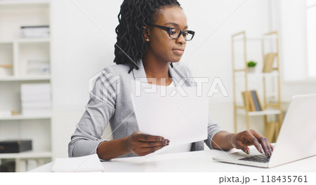 A focused black woman is diligently doing paperwork at her office desk. The mood is professional and organized, with a tidy workspace, modern decor, and a bright atmosphere, highlighting dedication. A focused black woman is diligently doing paperwork at her office desk. The mood is professional and organized, with a tidy workspace, modern decor, and a bright atmosphere, highlighting dedication. 118435761