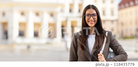 A confident young woman stands outside a historical building, wearing a fashionable outfit and glasses. Her warm smile suggests happiness on a bright, sunny day in the city. 118435779