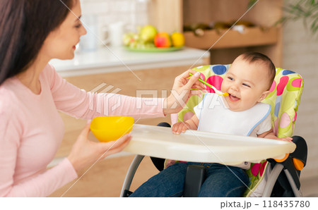 Beautiful woman sitting and feeding a baby with a spoon. The woman is holding the spoon while the baby is opening its mouth to eat. The setting seems to be indoors based on the background. 118435780