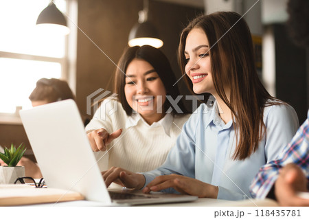 Two multiethnic women are seated at a desk in a well-lit library, working together on a project. One is pointing at a laptop screen, suggesting an idea or solution 118435781
