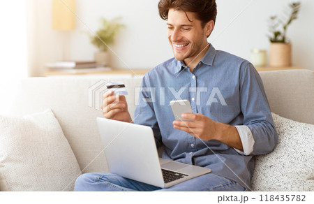 A man on a couch smiles while looking at his phone holding a credit card, with a laptop in front, implying hes making an online payment. 118435782
