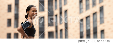 Fit Young Woman In Stylish Sportswear Resting Outdoors After Training, Sporty Athletic Lady With Earphones And Armband Phone Holder Standing On Urban City Street And Smiling At Camera, Copy Space 118435830