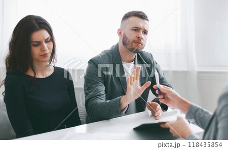 A couple sits at a table in a meeting with a lawyer, with the man gesturing and refusing to sign papers being handed to them. The woman sits beside the man, with a concerned expression on her face. A couple sits at a table in a meeting with a lawyer, with the man gesturing and refusing to sign papers being handed to them. The woman sits beside the man, with a concerned expression on her face. 118435854