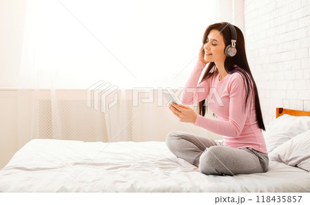 Young millennial woman relaxing and listening to music using wireless headphones, sitting in bed, empty space Young millennial woman relaxing and listening to music using wireless headphones, sitting in bed, empty space 118435857