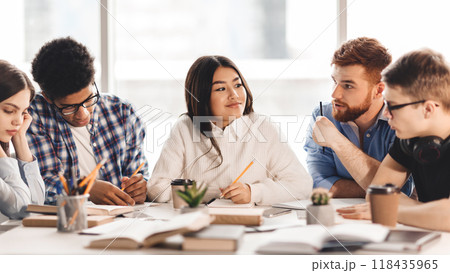 A diverse group of students sitting together at a table, engaged in conversation and possibly doing homework. Some are gesturing while speaking, while others are listening attentively A diverse group of students sitting together at a table, engaged in conversation and possibly doing homework. Some are gesturing while speaking, while others are listening attentively 118435965