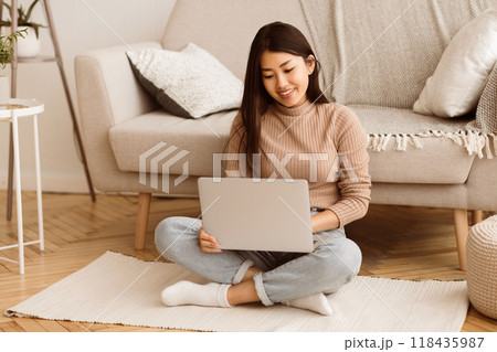 A woman sits on a rug in a living room, using a laptop computer. She is smiling and appears to be focused on her work. 118435987