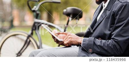 A businessman is sitting on a bench in a park, using a digital tablet with his bicycle close by. The atmosphere is tranquil and efficient, showing a modern work-life balance, cropped A businessman is sitting on a bench in a park, using a digital tablet with his bicycle close by. The atmosphere is tranquil and efficient, showing a modern work-life balance, cropped 118435998