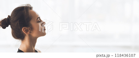 A young woman sits peacefully, eyes closed, in a well-lit room, practicing mindfulness and meditation, copy space 118436107